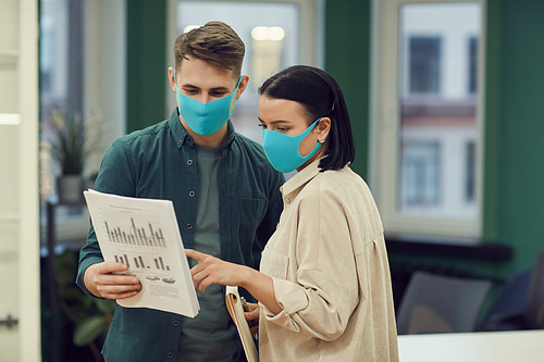 Two colleagues wearing protective masks standing at office and they discussing financial document