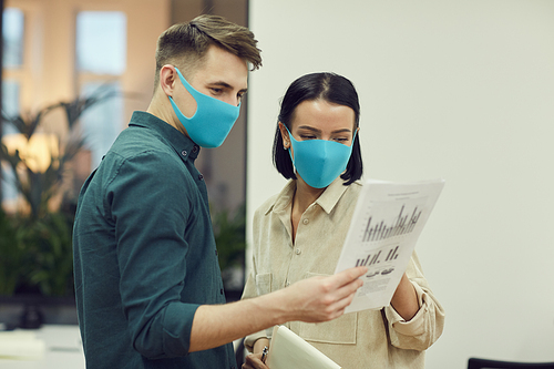 Young businessman in protective mask showing financial document to the businesswoman in protective mask and they examining it together