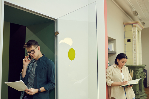 Young businessman holding documents and talking on the phone with businesswoman making notes in notepad in the background they working at office