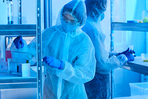 Asian young woman in protective workwear analysing chemicals in test tubes with her colleague working in the background at office