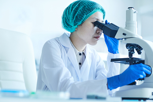 Asian young woman in white coat sitting at the table and examining analysis with microscope at office