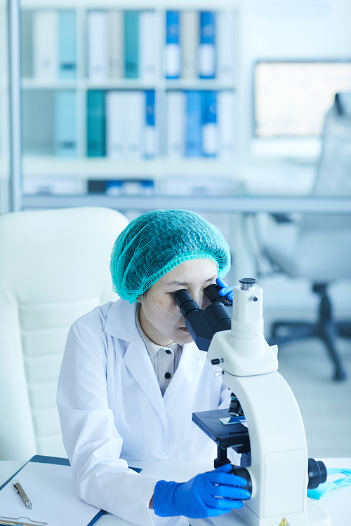 Female doctor in white coat sitting at the table and looking through the microscope and examining the samples at hospital