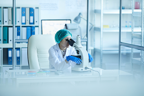 Young woman in white coat sitting at the table and looking through the microscope she working at her office