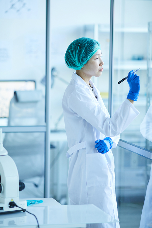Female scientist in white coat writing plans on glass wall while working at office