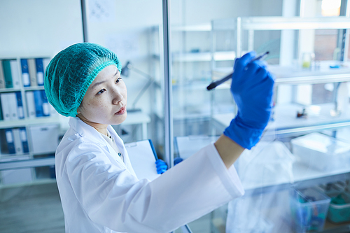 Asian female doctor in white coat and protective gloves making notes on glass wall at office