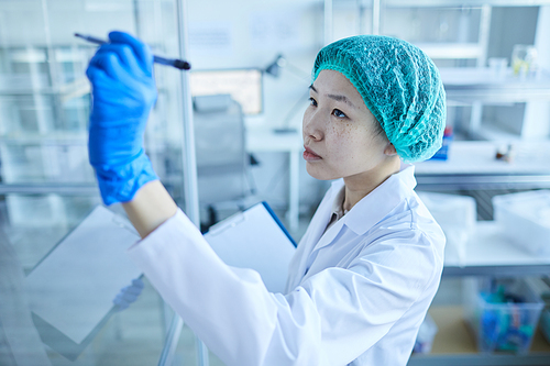Asian female doctor in white coat and in protective gloves and cap writing something on glass wall at hospital