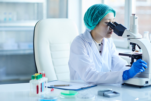 Female doctor sitting at the table and examining blood samples using microscope in her work at the lab