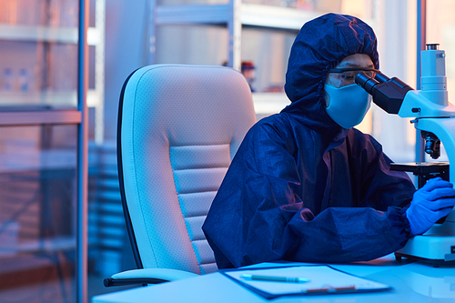 Doctor in protective workwear and in mask looking through the microscope while sitting at her workplace at office