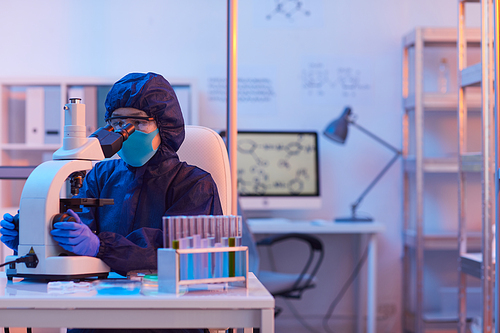 Scientist in protective workwear and mask sitting at her workplace with test tubes and examining samples through the microscope