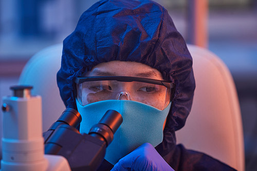 Close-up of female scientist in protective mask and glasses looking through the microscope while working at the lab