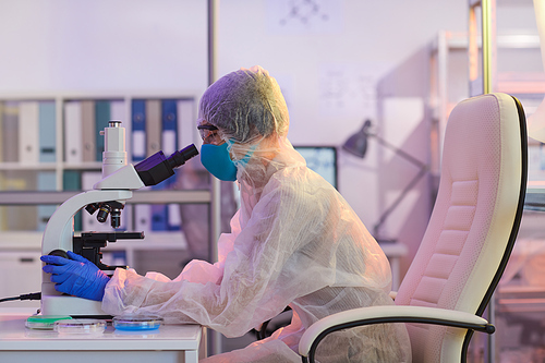 Medical worker in protective costume sitting at the table and looking through the microscope while working at the lab