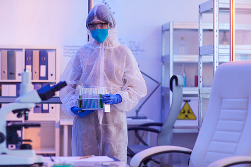Female chemist in protective costume holding test tubes and looking at camera while working in the laboratory