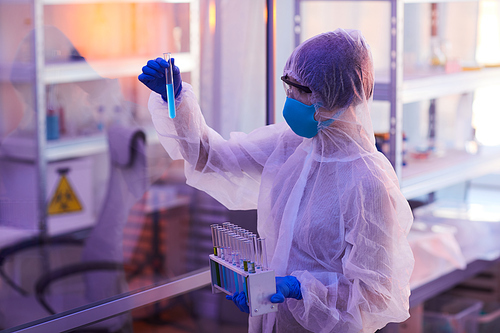 Scientist in protective workwear and in mask holding test tube with blue liquid and examining it in the lab
