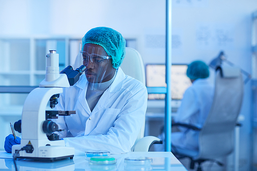 African young scientist sitting at his workplace and working with microscope during scientific experiment at office