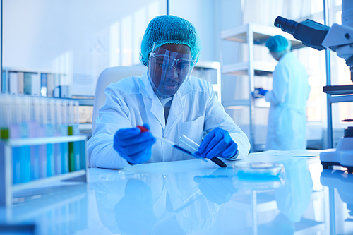 African scientist in white coat sitting at the table and working with samples in tubes