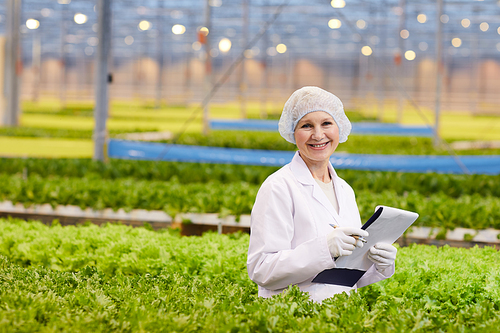 Portrait of mature woman in white lab coat holding document and smiling at camera while growing lettuce in greenhouse