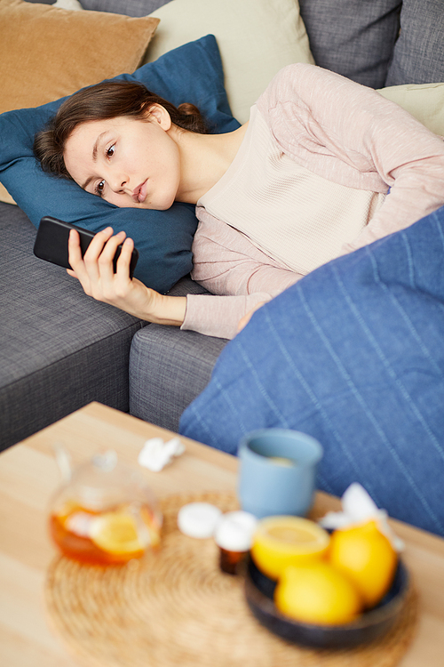 Young woman lying on the sofa drinking tea with lemon and using her mobile phone she feels herself unwell