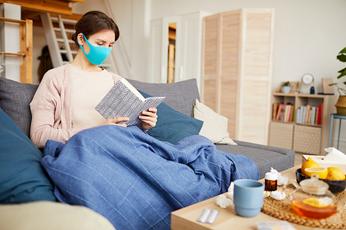 Young woman wearing protective mask resting on sofa and reading a book she is ill and sitting at home during quarantine