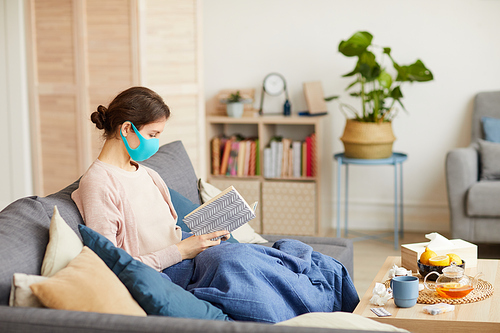 Sick woman wearing protective mask sitting on sofa with book she is on sick leave and sitting at home