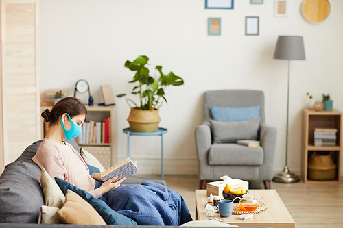 Young woman in protective mask sitting on quarantine she relaxing on sofa with book in the living room