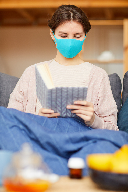 Young sick woman wearing protective mask sitting on sofa and reading a book at home