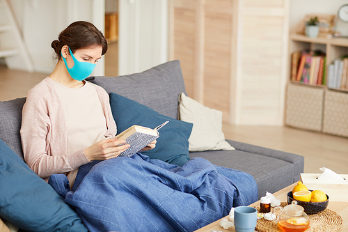 Young woman wearing protective mask she caught a cold and stay at home with book in the living room