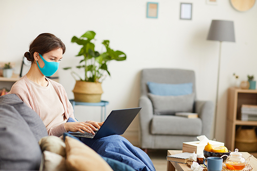 Young woman wearing protective mask sitting on sofa and typing on laptop computer she working online at home because of quarantine