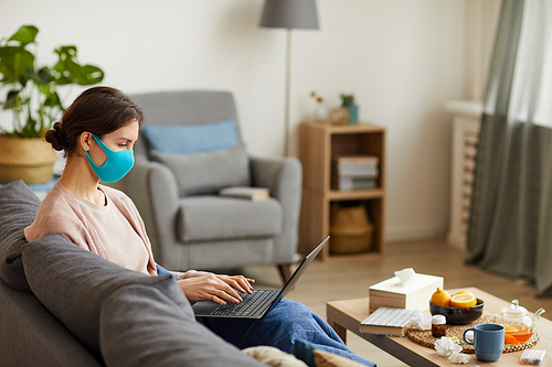 Young woman in protective mask working online on her laptop while sitting on sofa in the living room she stay at home because of pandemic