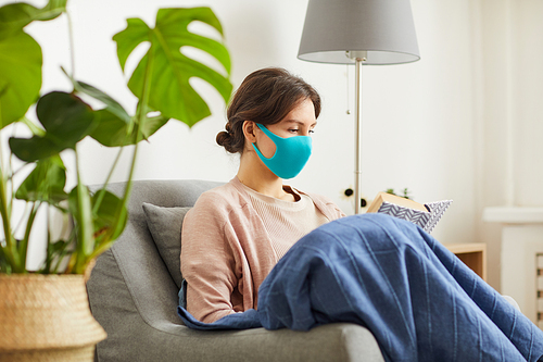 Young woman wearing protective mask sitting on armchair and reading her favourite book at home