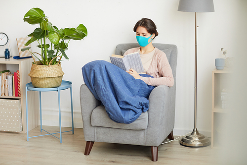 Young woman in protective mask sitting on quarantine she reading the book on armchair in the living room