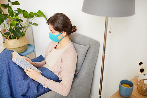 Young woman resting on armchair and reading a book at home she is ill and she wearing protective mask