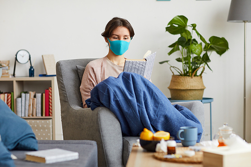 Young sick woman in protective mask sitting on armchair and reading a book she stay at home because of epidemic