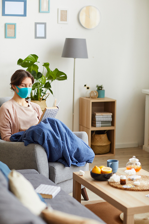 Sick woman in protective mask sitting at home on self isolation because of pandemic she reading a book in the living room