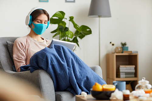 Young woman in protective mask and headphones sitting on armchair and using digital tablet she stay at home because of pandemic