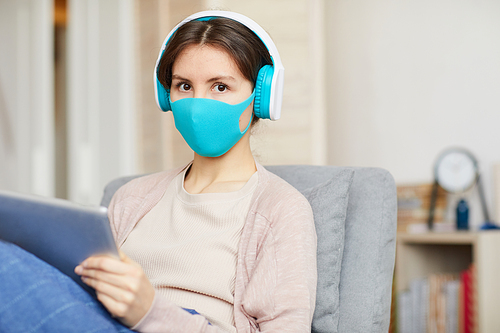 Portrait of young woman wearing headphones and protective mask looking at camera while listening to music on digital tablet