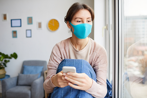 Young woman wearing protective mask she is ill and sitting at home she using her mobile phone and looking through the window