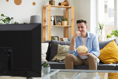 Young man sitting on sofa eating popcorn and watching his favourite film on TV at home