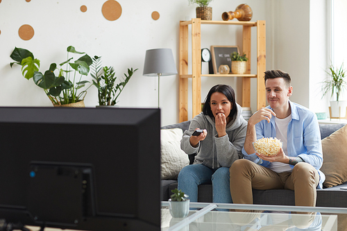 Young couple sitting on sofa eating popcorn and watching interesting film on TV at home