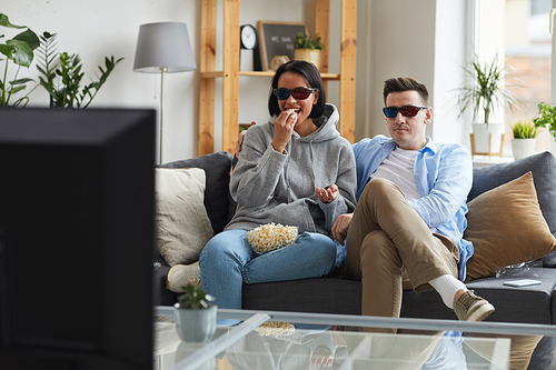 Happy young couple wearing 3D glasses sitting on sofa and eating popcorn while watching the movie on TV