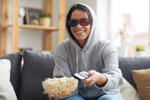 Smiling young woman in 3D glasses eating popcorn and watching TV on the sofa at home