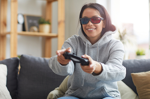 Young smiling woman in 3D glasses sitting on sofa with joystick and playing computer game at home
