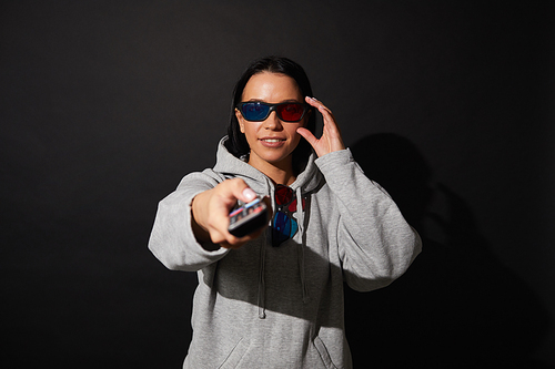 Portrait of young woman in 3D glasses holding remote control and switching on the TV over black background