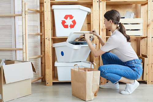 Young woman sitting near the trash bins and separating paper from other waste in warehouse