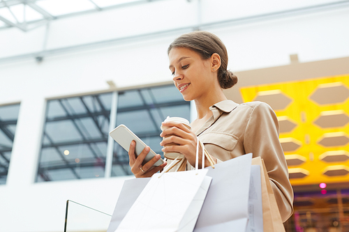 Young woman with shopping bags using mobile phone and drinking coffee during shopping in the store