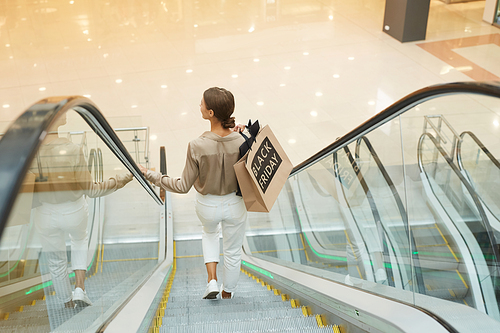 Rear view of young woman holding shopping bags and moving down on the escalator in shopping mall