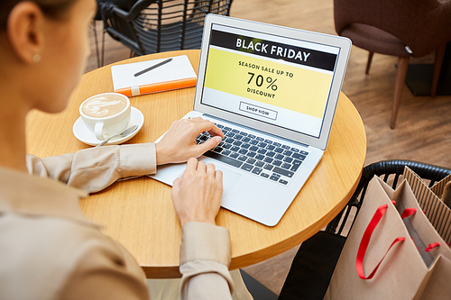 Rear view of young woman sitting at the table and using laptop computer she is going to buy purchases online in cafe