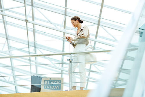 Young pretty woman using her mobile phone and communicating online while standing in modern shopping mall