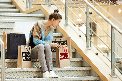 Young woman in casual clothing sitting on stairs and looking into the shopping bag after shopping in the shopping mall