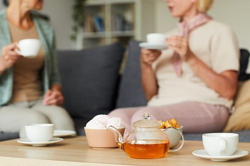 Close-up of teapot with tea with cups on the table and two women talking to each other on the sofa in the background