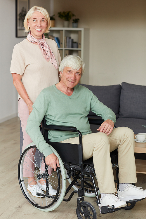 Portrait of senior woman smiling at camera while standing near her disabled husband in wheelchair